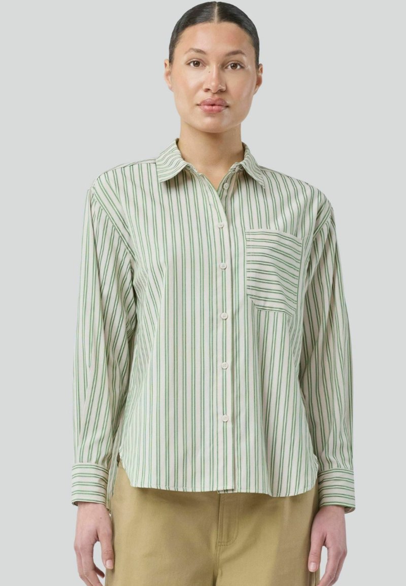 Modellen bærer en grøn og hvid stribet skjorte med knap, der har en brystlomme, og er iført beige bukser, mod en neutral grå baggrund.