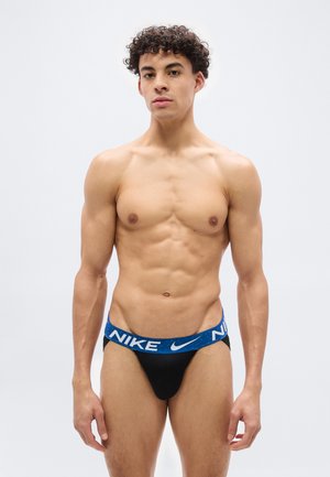 Jeune homme athlétique aux cheveux bouclés portant un slip noir Nike avec une ceinture bleue, debout devant un fond clair uni.