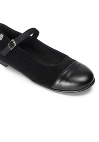 Zapato Mary Jane de ante negro con punta redonda y una tapa de cuero negro brillante. Presenta una correa delgada con una hebilla en tono plateado. Suela plana.