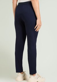 Donkerblauwe joggingbroek met een gladde textuur, elastische tailleband, taps toelopende pijpen en zijnaden. Draagt lichte sneakers.
