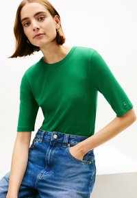 T-shirt verde a coste con maniche corte, scollatura rotonda, abbinato a jeans in denim blu a vita alta con tasca frontale e chiusura a bottoni.