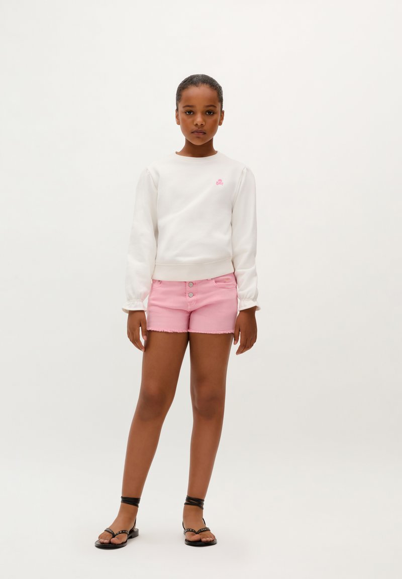 Weißer Sweatshirt mit Puffärmel, pinkem Logodetail. Pinke Jeans-Shorts mit fransigem Saum und drei Knöpfen. Schwarze Riemchensandalen.