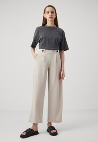T-shirt gris oversized, coupe décontractée. Pantalons larges crème avec fermeture à double bouton à la taille. Sandales plates noires avec des brides croisées.