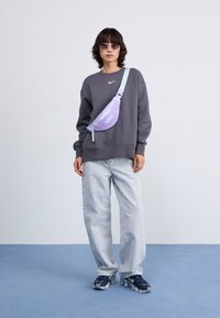 Transparante paarse heuptas met een lichtblauwe schouderband, gedragen over het lichaam; grijze sweatshirt en lichtblauwe jeans; tegen een minimalistische achtergrond.
