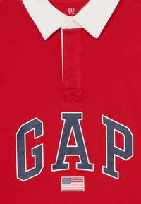 Rde majica polo z belim ovratnikom, velikimi mornarsko modrimi črkami "GAP" in majhno grafiko ameriške zastave spodaj. Mehka bombažna tkanina.