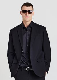 Schwarzer Anzug mit passendem Hemd, beide aus glattem Stoff. Der Look wird durch dunkle Sonnenbrillen und einen Gürtel mit silberner Schnalle vervollständigt.