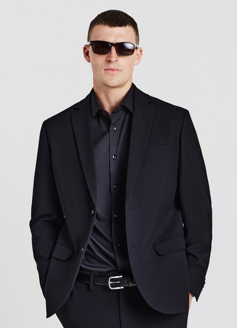 Schwarzer Anzug mit passendem Hemd, beide aus glattem Stoff. Der Look wird durch dunkle Sonnenbrillen und einen Gürtel mit silberner Schnalle vervollständigt.