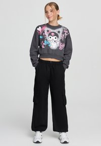 Felpa grigia corta con grafiche di cartoni animati in rosa e blu, abbinata a pantaloni cargo neri larghi con tasche laterali.