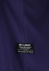 Tela morada con un patrón texturizado y una etiqueta cosida que dice "KEY LARGO", junto con información de marca y cuidado en texto blanco.