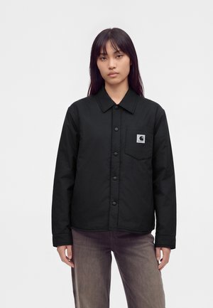 Femme aux cheveux raides et foncés portant une veste noire boutonnée avec une poche poitrine et un écusson logo sur un fond blanc uni.