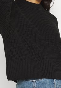 Svart stickad tröja med ribbat halsband och texturerade mönster, med en avslappnad passform och en mjuk yta. Materialet verkar mjukt och varmt.