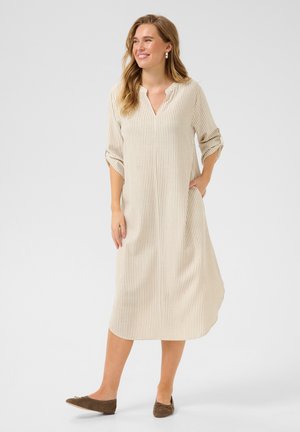 Striped beige dress with a V-neck, long sleeves rolled up, side pockets, and a loose fit. Paired with brown flat shoes. Smooth fabric texture.