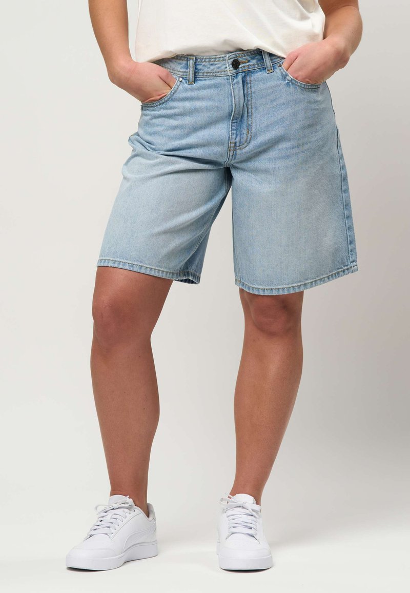 Personne portant un short en jean bleu clair jusqu'aux genoux, les mains dans les poches et des baskets blanches, debout devant un fond uni.