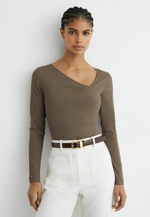 Frau mit geflochtenem Haar trägt ein eng anliegendes, olivgrünes Langarm-V-Neck-Oberteil, das in eine hoch taillierte weiße Hose mit dunkelbraunem Gürtel gesteckt ist.