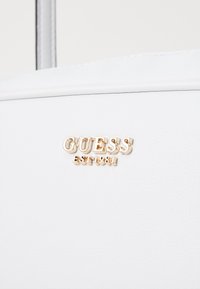 Witte synthetische handtas met een gladde textuur. Kenmerkt zich door een gouden "GUESS" logo en "EST 1981" detail, wat contrast toevoegt aan het minimalistische ontwerp.
