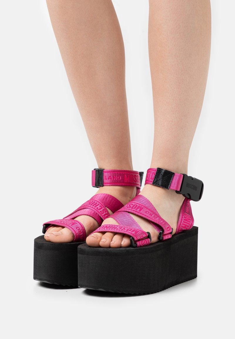 Pieds portant des sandales à plateforme noires avec des brides réglables rose vif et des boucles noires sur fond blanc.