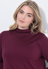 Burgundská roláková halenka s přiléhavým střihem, vyrobená z hladkého materiálu. S mírným leskem a dlouhými rukávy.