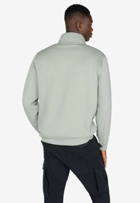 Helles graues Fleecepullover mit hohem Kragen und gerippten Bündchen, kombiniert mit dunklen Cargo-Hosen, die mehrere Taschen und strukturierte Akzente aufweisen.