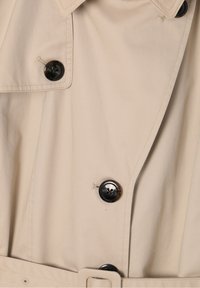 Trench-coat beige avec des boutons ronds foncés, un large col et une ceinture en tissu assortie à la taille.