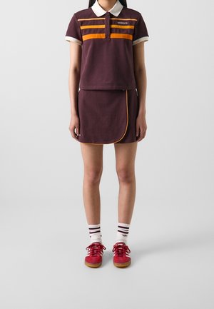Osoba ubrana w brązową koszulkę polo Adidas z pomarańczowymi paskami, dopasowaną spódnicę z pomarańczowym wykończeniem, białe skarpetki w paski i czerwone trampki.