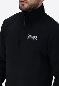 Czarny polarowy sweter z wysokim kołnierzem i zamkiem błyskawicznym na ćwierć długości z przodu. Haftowane białe logo "Lonsdale London" na piersi.