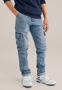 Gai zila džinsa bikses ar divām sānu kabatām, sašaurināta piegrieze, nedaudz izbalējusi tekstūra, kombinētas ar baltiem un jūras zilas krāsas sporta apaviem.