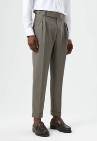 Pantalon plissé kaki avec une taille haute, des ajusteurs latéraux et des revers roulés, associé à des mocassins marron foncé et une chemise blanche.