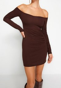 Hnědress s odhalenými rameny vyrobený z pružného materiálu, který má dlouhé rukávy a boční nařasení pro vybroušený siluetu.