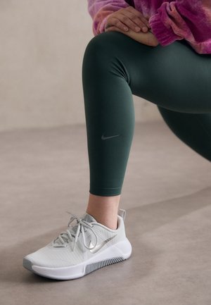 Persona con mallas verdes y camiseta rosa hace una zancada, con zapatillas blancas Nike en un suelo interior liso. Las manos descansan sobre la rodilla doblada.