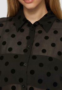 Zwarte sheer shirt met een stippenpatroon met textuur in zwarte stippen, een puntige kraag en knoopsluiting aan de voorkant.