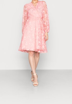Femme portant une robe portefeuille fleurie rose midi à manches longues et des talons beiges à bride cheville, debout devant un fond uni.