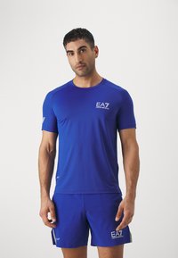 Camiseta deportiva azul, hecha de una tela elástica, con un escote redondo y mangas cortas. Logo EA7 en el pecho y en la manga.