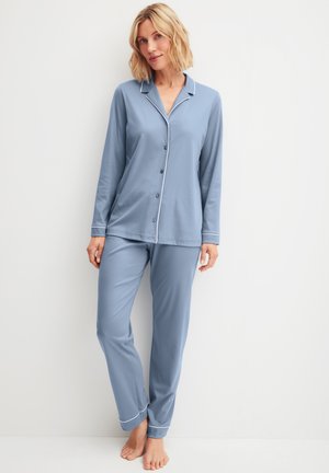 Vrouw met blond haar, gekleed in een lichtblauwe pyjamabovenkant met lange mouwen en knopen, en bijpassende broek, staand op blote voeten tegen een witte muur.