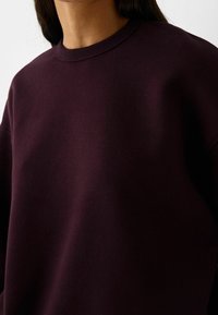 Persona vistiendo un suéter de cuello redondo de color púrpura oscuro con mangas largas, mostrado desde los hombros hasta la parte superior del pecho sobre un fondo liso.