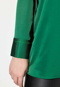 Zielona satynowa koszula o błyszczącej fakturze, z mankietem ozdobionym współrzędnymi w jasnej zieleni. Koszula ma gładki, płynny krój.