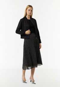 Czarna kurtka bombowa z prążkowanymi mankietami, zestawiona z plisowaną czarną spódnicą z metalicznymi kwiatowymi wzorami. Modelka nosi srebrne szpilki.