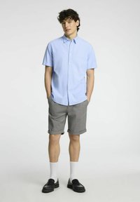 Camicia azzurro chiaro a maniche corte, pantaloni corti grigi con risvolti, mocassini neri e calze bianche. Stile casual, vestibilità rilassata, tessuto morbido.