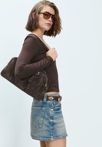 Rjav dolg rokav majica, denim mini krilo in teksturirana temno rjava torbica z zipperji. Model nosi sončna očala in ima kratke lase.