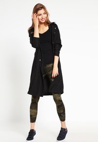 Black long cardigan with golden buttons, worn over a black top and green camo leggings. Features a relaxed fit and smooth fabric texture.