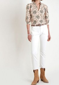 Hedvábná blůza s květinovým vzorem v broskvové barvě, s nabíranými rukávy, doplněná o bílé přiléhavé kalhoty a světle hnědé semišové kotníkové boty s dekorativním designem.