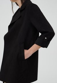 Femme portant un manteau noir aux manches retroussées, la main dans la poche latérale, vue de profil sur un fond uni.