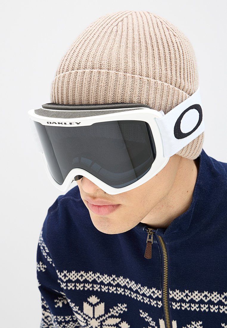 Occhiali da sci bianchi con lenti scure, imbottitura in schiuma nera e fascia regolabile con logo "Oakley". Il modello indossa un berretto a maglia beige e un maglione decorato.