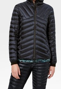 Schwarze Steppjacke mit Zickzack-Steppung, hohem Kragen und durchgehendem Reißverschluss. Glatte, glänzende Oberfläche mit elastischen Bündchen und Saum. Kombiniert mit passenden schwarzen Hosen.