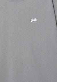 Grå bomuld t-shirt med et lille hvidt broderet logo på venstre brystområde. Stoffet har en glat, ensartet tekstur.