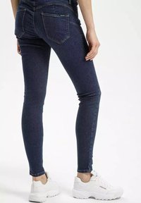 Cienkie dżinsy w ciemnoniebieskim deninie z wyblakłymi akcentami, z pięcioma kieszeniami i dopasowanym krojem. Noszone z białymi sportowymi sneakersami.