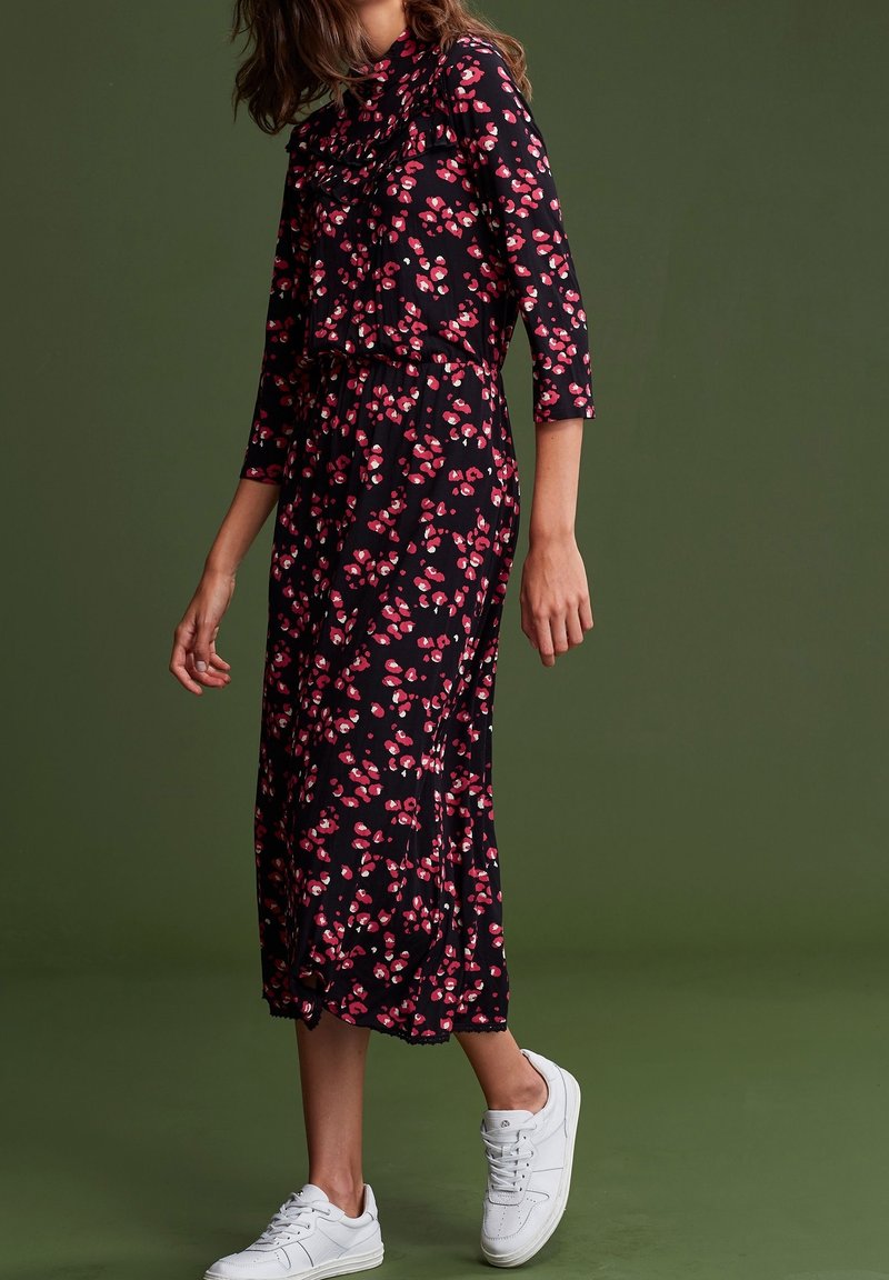 Femme portant une robe midi noire à imprimé floral rose avec des manches trois-quarts, associée à des baskets blanches, debout devant un fond vert.