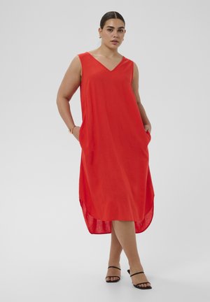 Žena v červenom midi šatách bez rukávov a s výstrihom do V, so započenými vreckami, stojaca so skríženými nohami, v čiernych sandáloch na podpätku a zlatých kruhových náušniciach.