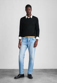 Μαύρος σστόσοερ πάνω από άσπρο πουκάμισο με γιακά, ανοιχτό μπλε τζιν παντελόνι, μπεζ ζώνη, μαύρα παπούτσια, σ standing απέναντι σε απλό ανοιχτό γκρί φόντο.