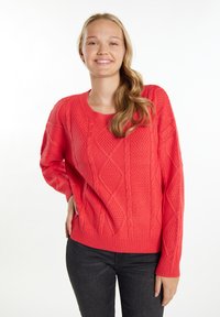 Koralowy sweter dzianinowy z teksturowanymi wzorami w diamenty i warkocze, okrągły dekolt i ściągacze na rękawach, w połączeniu z ciemnymi dżinsami.