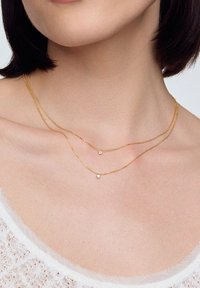 Złototona, warstwowa naszyjnik z cienkim łańcuszkiem i dwoma małymi, błyszczącymi akcentami, odpoczywającymi w okolicy obojczyka. Gładka tekstura, delikatny design.